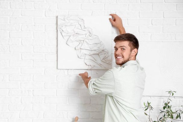 Молодой бородатый мужчина висит картину на белой кирпичной стене дома - Фото, изображение