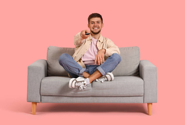 Jeune homme barbu regardant la télévision sur un canapé gris sur fond rose - Photo, image