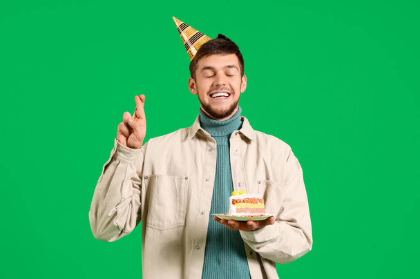 Jeune homme barbu en chapeau de fête avec morceau de gâteau d'anniversaire faire souhait sur fond vert - Photo, image