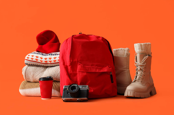 Mochila con ropa de abrigo, cámara y taza de café sobre fondo naranja. concepto de vacaciones de invierno - Foto, Imagen
