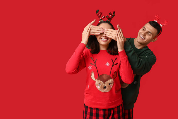 Happy young man in Christmas pajamas covering eyes of wife with his hands on red background - Photo, Image