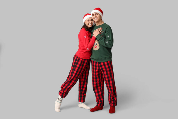 Noel pijamaları ve gri arka planda Noel Baba şapkaları giyen mutlu çift. - Fotoğraf, Görsel