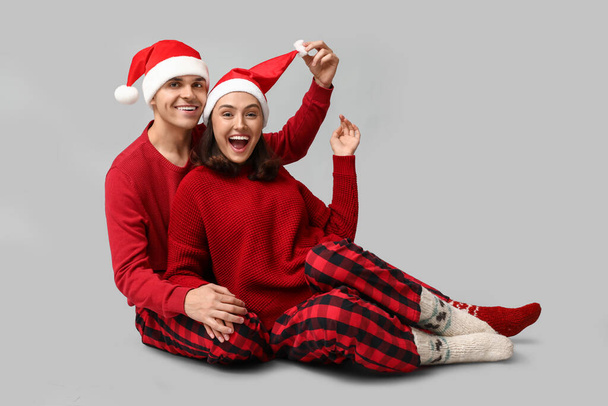 Happy young couple in Christmas pajamas and Santa hats sitting against grey background - Photo, Image