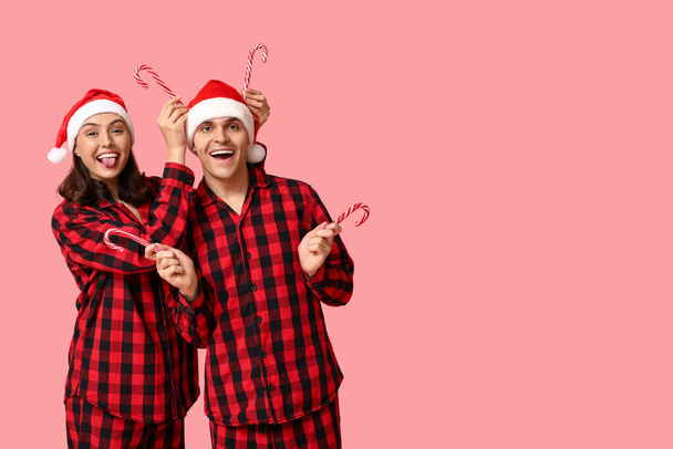 Hauska nuori pari joulupyjamassa ja karkkitangoilla vaaleanpunaisella taustalla - Valokuva, kuva
