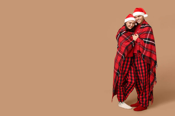 Happy young couple in Christmas pajamas and with warm plaid hugging on beige background - Photo, Image
