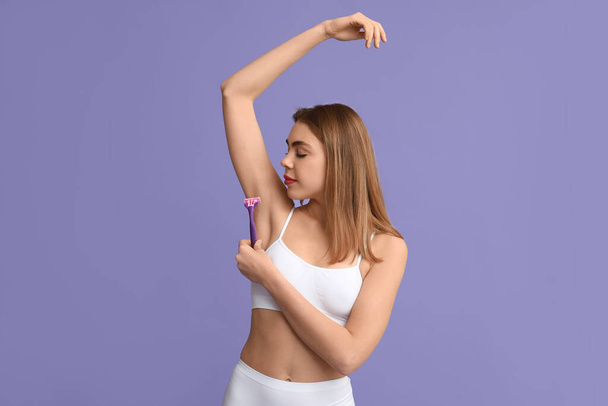 Beautiful young woman with razor shaving armpit on purple background - Photo, Image