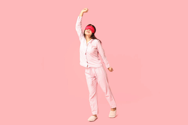 Hermosa joven asiática en pijama con máscara de dormir sobre fondo rosa - Foto, Imagen