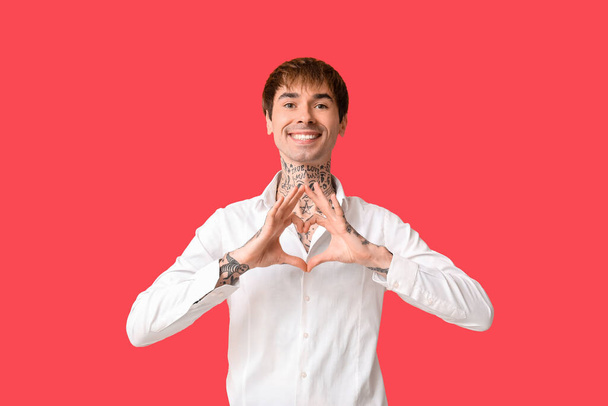 Ευτυχισμένος όμορφος νεαρός άνδρας με τατουάζ που δείχνει χειρονομία καρδιάς σε κόκκινο φόντο - Φωτογραφία, εικόνα