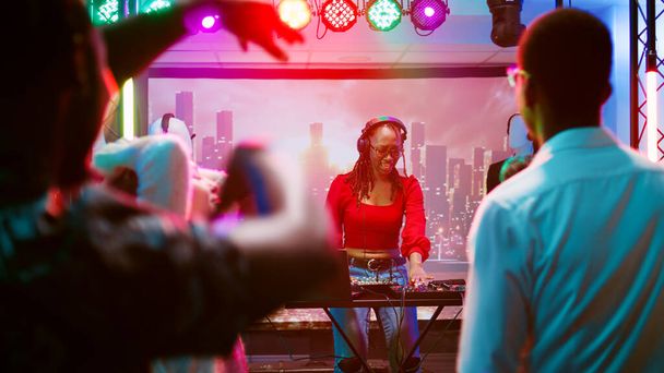 女性DJがナイトクラブで音楽をミックスし、ダンスフロアの多様なグループとパーティーします。ディスコパーティーでディスコを楽しんでいるファンキーな女の子、クラブステージ上のオーディオステーション。手持ち撮影. - 写真・画像