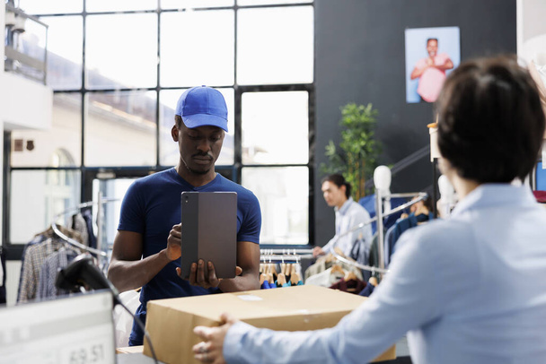 Afrikanisch-amerikanischer Kurier fotografiert Angestellte mit Tablet-Computer, Ladenangestellte hält Pappschachtel in moderner Boutique. Mitarbeiter eines Einkaufszentrums bereitet Pakete für die Zustellung vor - Foto, Bild