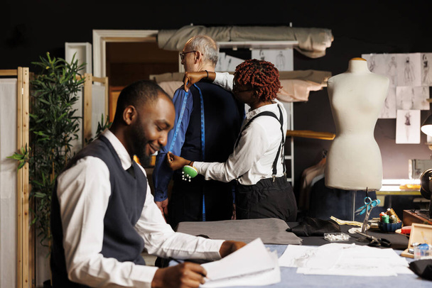 Teamworking couturiers helping elderly customer find perfectly fitting bespoke business suit. Happy tailors taking senior client measurements during sartorial customization comission process - Photo, Image