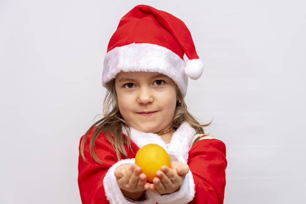kleines Mädchen als Weihnachtsmann verkleidet hält eine Orange auf weißem Hintergrund in die Höhe - Foto, Bild