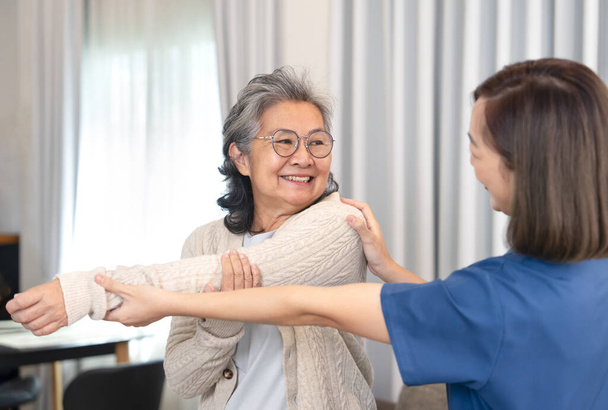positive Emotionen Seniorin, die Physiotherapie macht, die von einer jungen Krankenschwester unterstützt wird, asiatischer älterer Erwachsener mit Reha-Armdehnung zu Hause, Konzept des Lebensstils älterer Menschen, häusliche Pflege, häusliche Krankenpflege - Foto, Bild