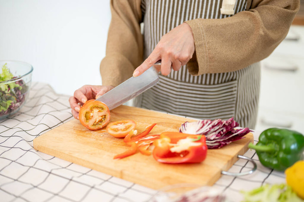 Close-up image of a woman in an apron is chopping a tomato, preparing fresh ingredients for her food in the kitchen. Home cooking, healthy food, diet meal - Photo, Image