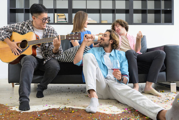 Groep multi-etnische vrienden die samen plezier hebben terwijl ze thuis op de bank zitten. man in bronde haar zingen een lied terwijl aziatische vrouw spelen gitaar in woonkamer. - Foto, afbeelding