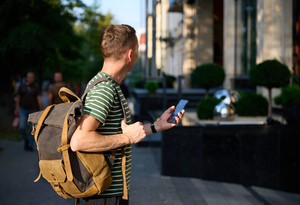Fiatal sportos férfi csíkos pólóban sétál az utcán okostelefonnal a kezében és egy divatos hipszter hátizsákja a válla fölött, élvezve egy kényelmes városi kirándulást.. - Fotó, kép