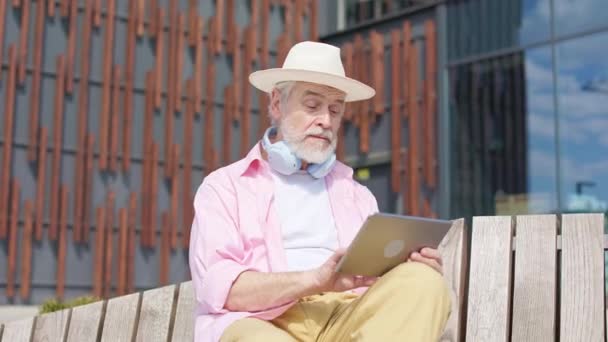 Zelfverzekerde oudere man zittend op houten bank en gericht op digitale tablet in handen. Serieuze blanke heer met behulp van moderne gadgets terwijl u geniet van vrije tijd in de buitenlucht. - Video
