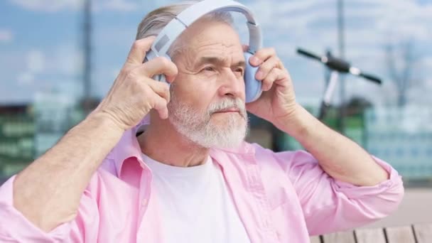 Πορτρέτο του ηλικιωμένου άνδρα σε άνετη ενδυμασία φορώντας ασύρματα ακουστικά, ενώ κάθεται σε ξύλινο πάγκο στο ηλιόλουστο κέντρο της πόλης. Καυκάσιος άνδρας απολαμβάνει τη μουσική και κοιτάζει προσεκτικά την κάμερα. - Πλάνα, βίντεο