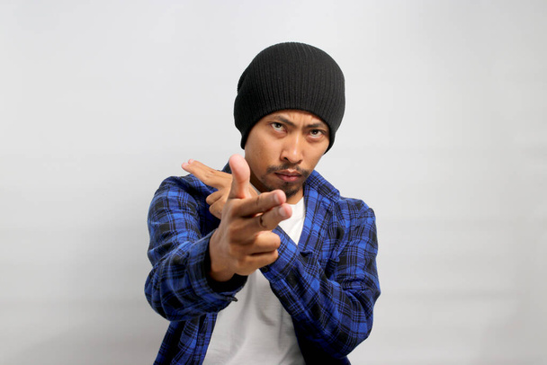 Confident young Asian man, points at the camera with a finger gun gesture, displaying confidence, making choices, and aiming with a hand pistol gesture right on target while standing on white backdrop - Photo, Image