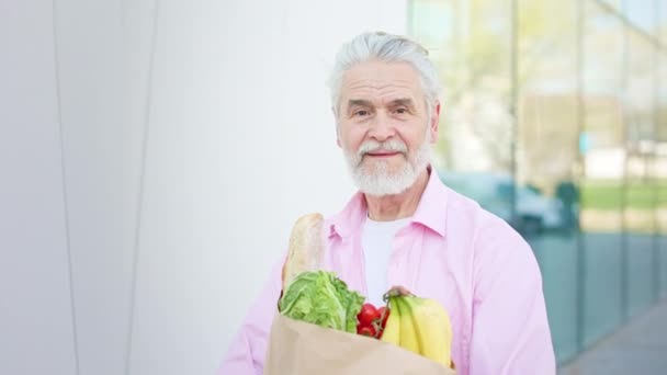 Portret przystojnego białego brodatego mężczyzny trzymającego odżywczy produkt w opakowaniu rzemiosła na zewnątrz. Starszy siwowłosy mężczyzna ubrany w luźną różową koszulę uśmiechnięty i patrzący w kamerę. - Materiał filmowy, wideo
