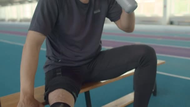 Tilt up shot van Kaukasische mannelijke para-atleet met ledematen prothese zittend op bank in indoor stadion en drinkwater, rusten na vermoeiende training - Video