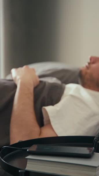 Вертикальный боковой удар бородатого кавказца, который просыпается от тревоги, когда он заряжает телефон и встает с кровати дома - Кадры, видео