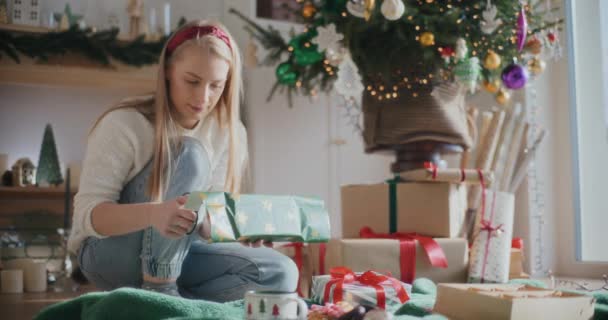 Όμορφη νεαρή γυναίκα κοπής χαρτιού, ενώ περιτύλιγμα χριστουγεννιάτικο δώρο στο σπίτι - Πλάνα, βίντεο