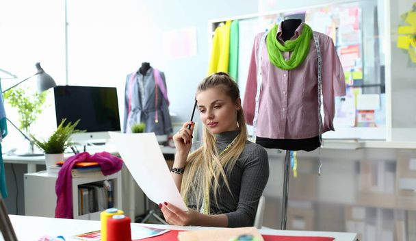 服のデザインプロセスと縫製コンセプトの作成ドレススケッチに取り組む美しいサンプストレス思考。現代のドレスメイキングワークショップに座っているペンシブ白人女性。仕立てビジネス - 写真・画像
