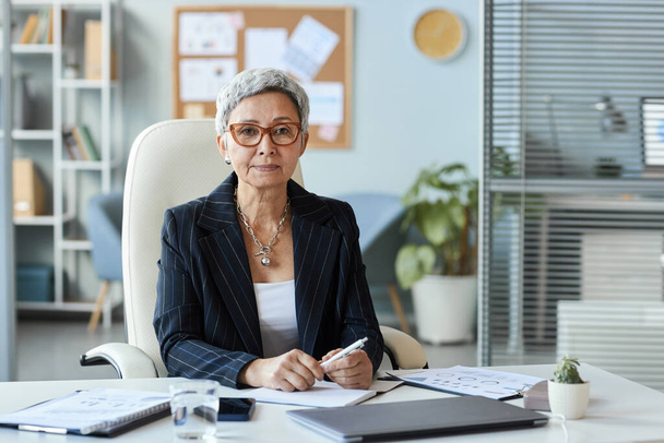 Porträt einer Seniorin als weibliche Chefin, die am Arbeitsplatz im Büro sitzt und in die Kamera schaut, Kopierraum - Foto, Bild