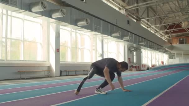 Длинный кавказский спортсмен садится на крытый трек и снимает протез с ампутированной конечности после завершения гонки на стадионе - Кадры, видео
