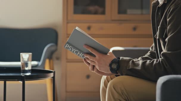 Corte lado esfaqueamento imagens de homem irreconhecível lendo livro psicologia sentado no sofá e sair depois de verificar o tempo de relógio na sala de estar moderna - Filmagem, Vídeo
