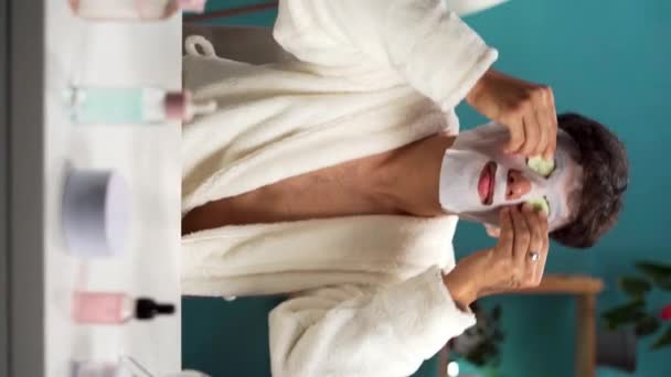 Handsome young man with facial mask on his face putting slices of fresh cucumber on eyes. Skin care, treatment and cosmetology concept. Vertical video - Footage, Video