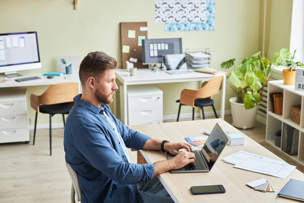 Πλάγια άποψη πορτρέτο του γενειοφόρου νεαρός άνδρας χρησιμοποιώντας φορητό υπολογιστή, ενώ εργάζονται σε άνετο γραφείο, αντίγραφο χώρο - Φωτογραφία, εικόνα
