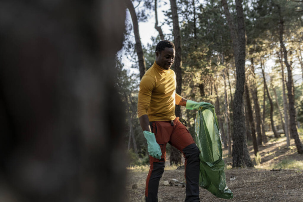 Afrikkalainen amerikkalainen vapaaehtoinen muovipussin kanssa kerää jätteitä ja roskia metsässä - Valokuva, kuva