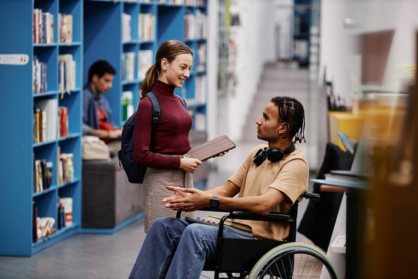 Портрет молодого студента з інвалідністю, що розмовляє з другом у барвистій бібліотеці коледжу, простір для копіювання - Фото, зображення