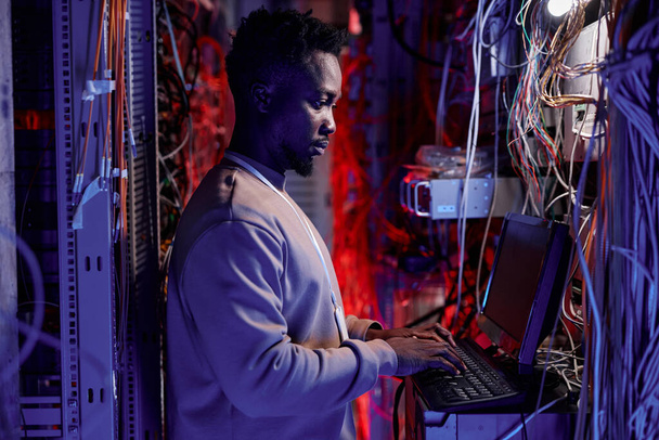 Πλάγια απεικόνιση του μαύρου ανθρώπου ως διαχειριστή δικτύου που δημιουργεί διακομιστές και χρησιμοποιεί φορητό υπολογιστή σε νέον φως - Φωτογραφία, εικόνα