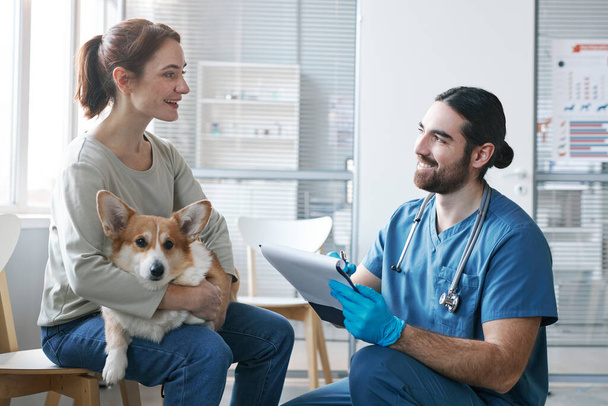Young male veterinarian with medical document talking to owner of purebred welsh pembroke corgi dog during consultation - Photo, Image