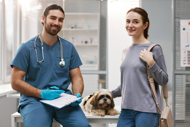 Mladá fena majitel roztomilý yorkshire teriér, její mazlíček a veterinář klinik v modré uniformě při pohledu na kameru v lékařské ordinaci - Fotografie, Obrázek