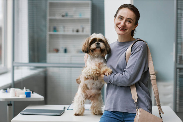 Onnellinen nuori naispuolinen omistaja söpö yorkshire terrieri tilalla hänen pörröinen lemmikki ja katsot kameraa istuessaan lääketieteellisessä toimistossa eläinlääkärin klinikat - Valokuva, kuva