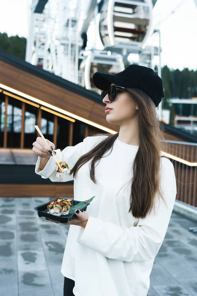 A pretty woman in a black cap eats sushi against the background of the Ferris wheel and the mountains. Sushi concept - Fotó, kép