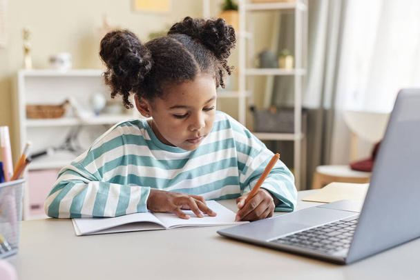 Porträt eines süßen schwarzen Mädchens, das in Notizbuch schreibt, während es Hausaufgaben am Schreibtisch in minimalem Home Interieur macht - Foto, Bild