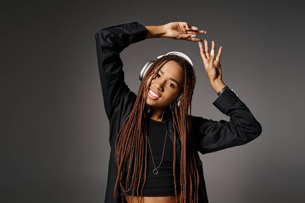 mujer afroamericana feliz y joven de 20 años con auriculares bailando al son de la música sobre fondo gris - Foto, Imagen