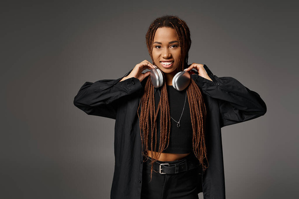 smiling african american woman with dreadlocks standing in headphones and looking at camera on gray - Photo, Image