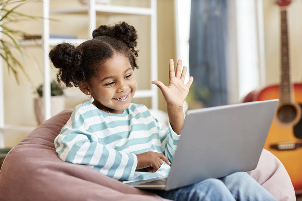 Küçük siyah kızın portresi dizüstü bilgisayara el sallıyor ve gülümsüyor. Evde rahat bir fasulye koltukta oturmuş, fotokopi çekerken çevrimiçi dersten keyif alıyor. - Fotoğraf, Görsel
