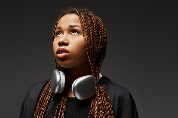 ονειρεμένη Αφροαμερικανή γυναίκα με κοτσιδάκια να στέκεται με ακουστικά και να κοιτάει πάνω στο γκρι. - Φωτογραφία, εικόνα
