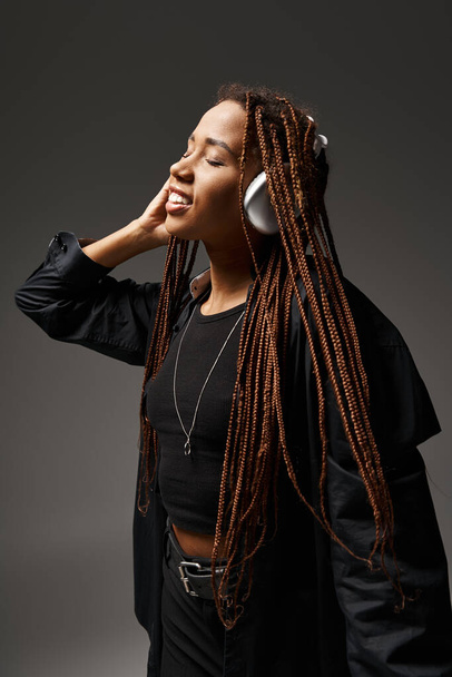 positive african american girl in 20s with dreadlocks listening music in headphones on grey backdrop - Photo, Image