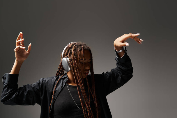 ecstatic dance of african american girl in her 20s with dreadlocks in wireless headphones on grey - Photo, Image