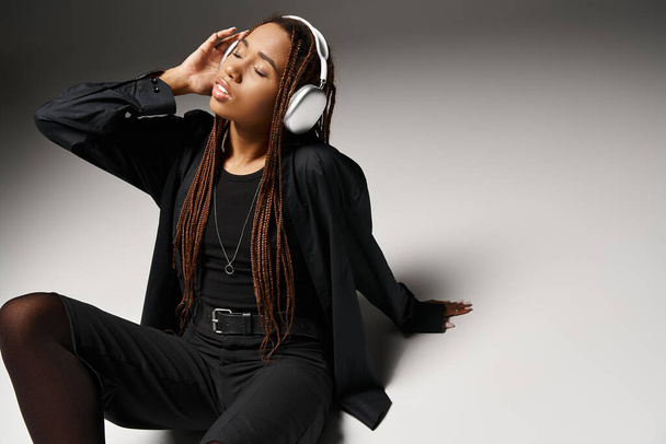 Αφροαμερικανή 20άρα με κλειστά μάτια που απολαμβάνει μουσική με ακουστικά και κάθεται πάνω σε γκρι. - Φωτογραφία, εικόνα