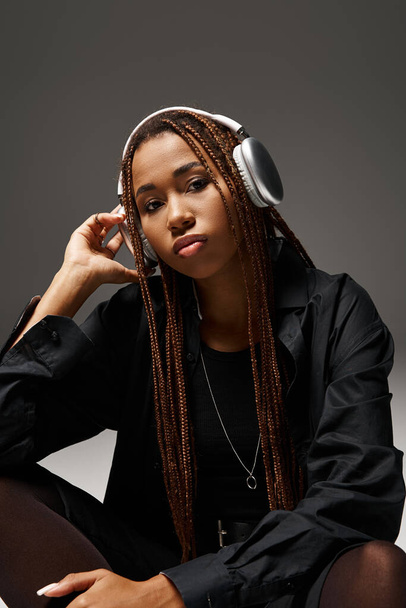 young african american woman in 20s with dreadlocks listening music in stylish headphones on grey - Photo, Image