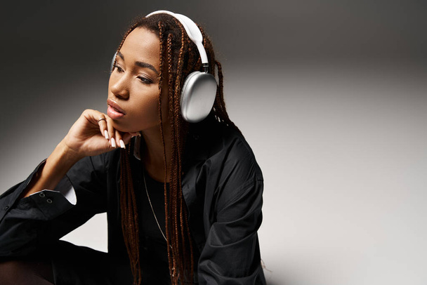 Pensive αφροαμερικανή γυναίκα στα 20 με dreadlocks ακούγοντας μουσική σε ακουστικά σε γκρι στούντιο - Φωτογραφία, εικόνα
